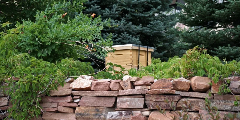 A wooden beehive nestled in a lush garden with robust stone wall and evergreen trees