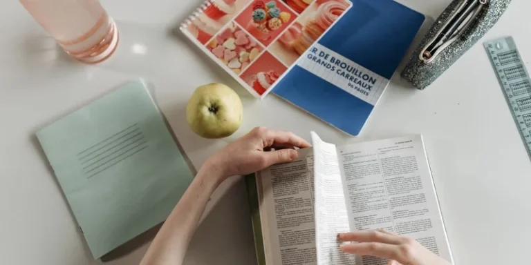 Overhead view of hands turning book pages at a study desk with notebooks, apple, and water bottle