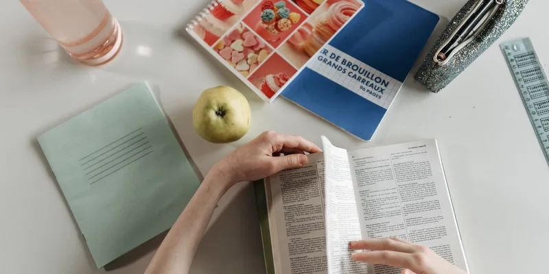 Overhead view of hands turning book pages at a study desk with notebooks, apple, and water bottle