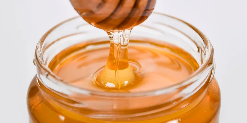 Close-up of honey flowing from a wooden dipper into a glass jar on a white background