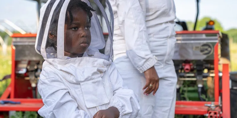 A young child in protective beekeeping clothing stands outdoors during a sunny day