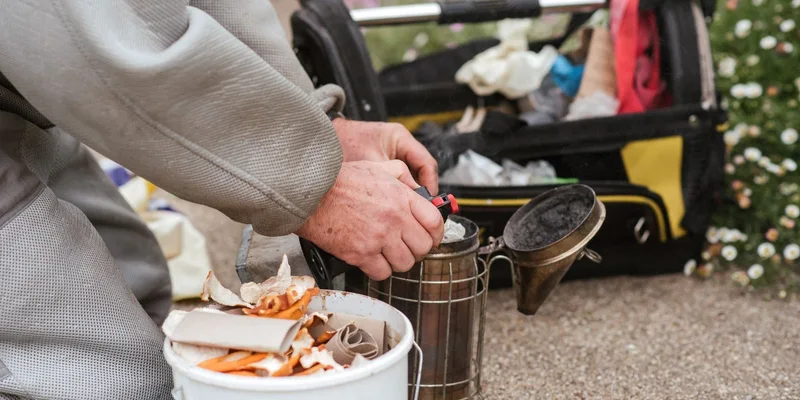 A beekeeper prepares a bee smoker outdoors, surrounded by equipment and nature